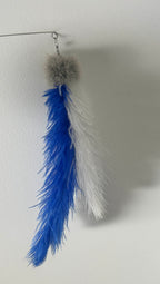 A hanging feather toy with two feathers, one bright blue and one white, attached to a gray fuzzy pom-pom top, suspended from a thin wire against a plain light wall.