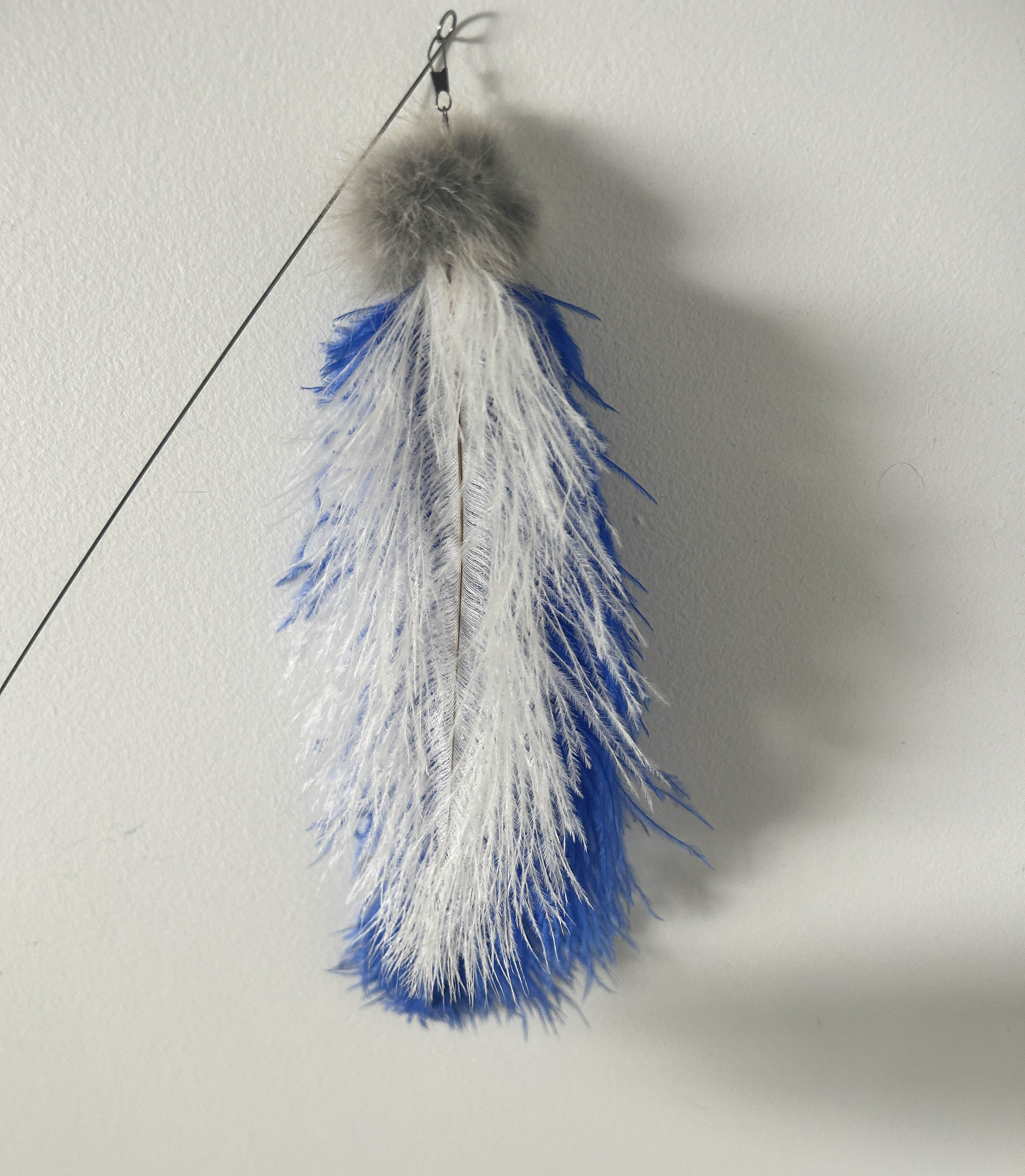 A blue and white feather toy with a gray fluffy pom-pom top and metal hanging clip, shown against a plain off-white wall.