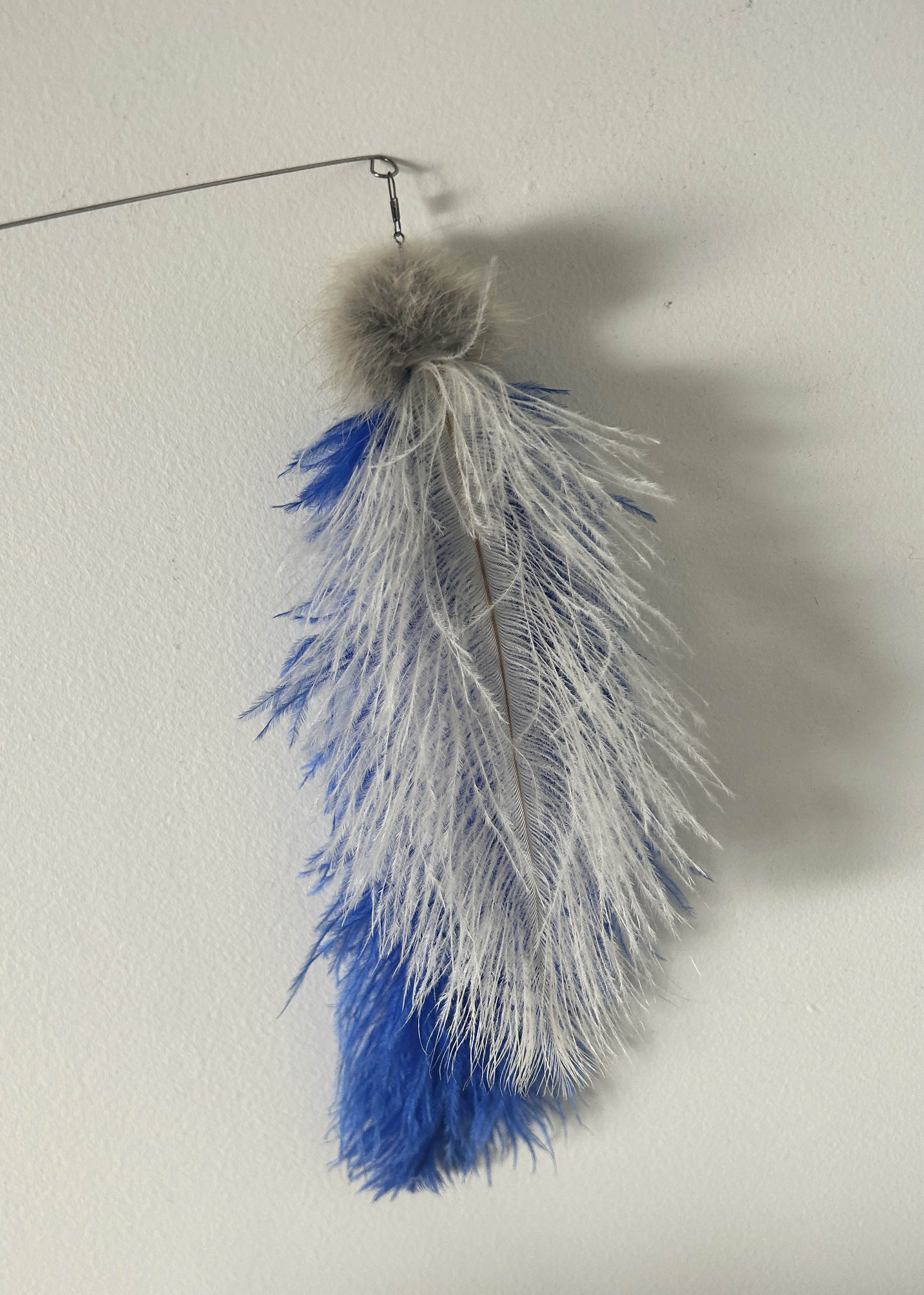 A decorative hanging feather cat toy with a gray faux-fur top, a central white feather, and blue feather accents, suspended from a thin wire against a plain off-white wall.