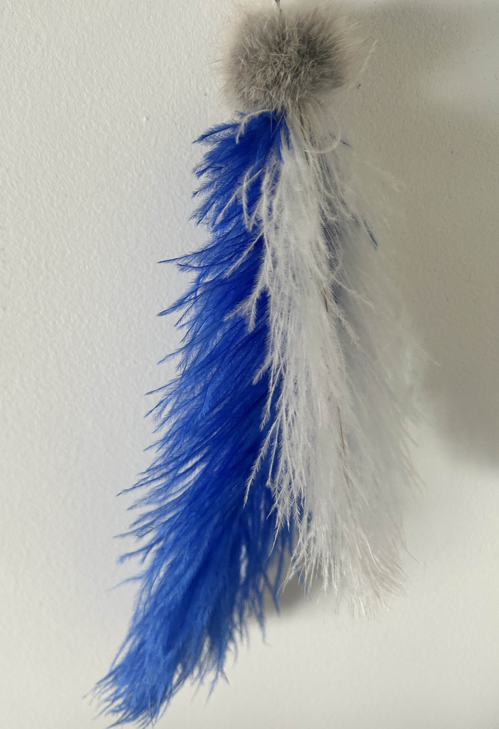 A cat toy consisting of a gray faux fur pom-pom attached to two feather plumes, one bright blue and one white, shown against a plain off-white wall.