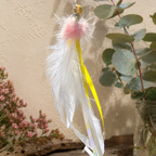 A decorative ribbon cat toy with a fluffy pink pom-pom, white and yellow feathers, and a gold ring, photographed against a textured beige wall with eucalyptus leaves and small pink flowers as props.
