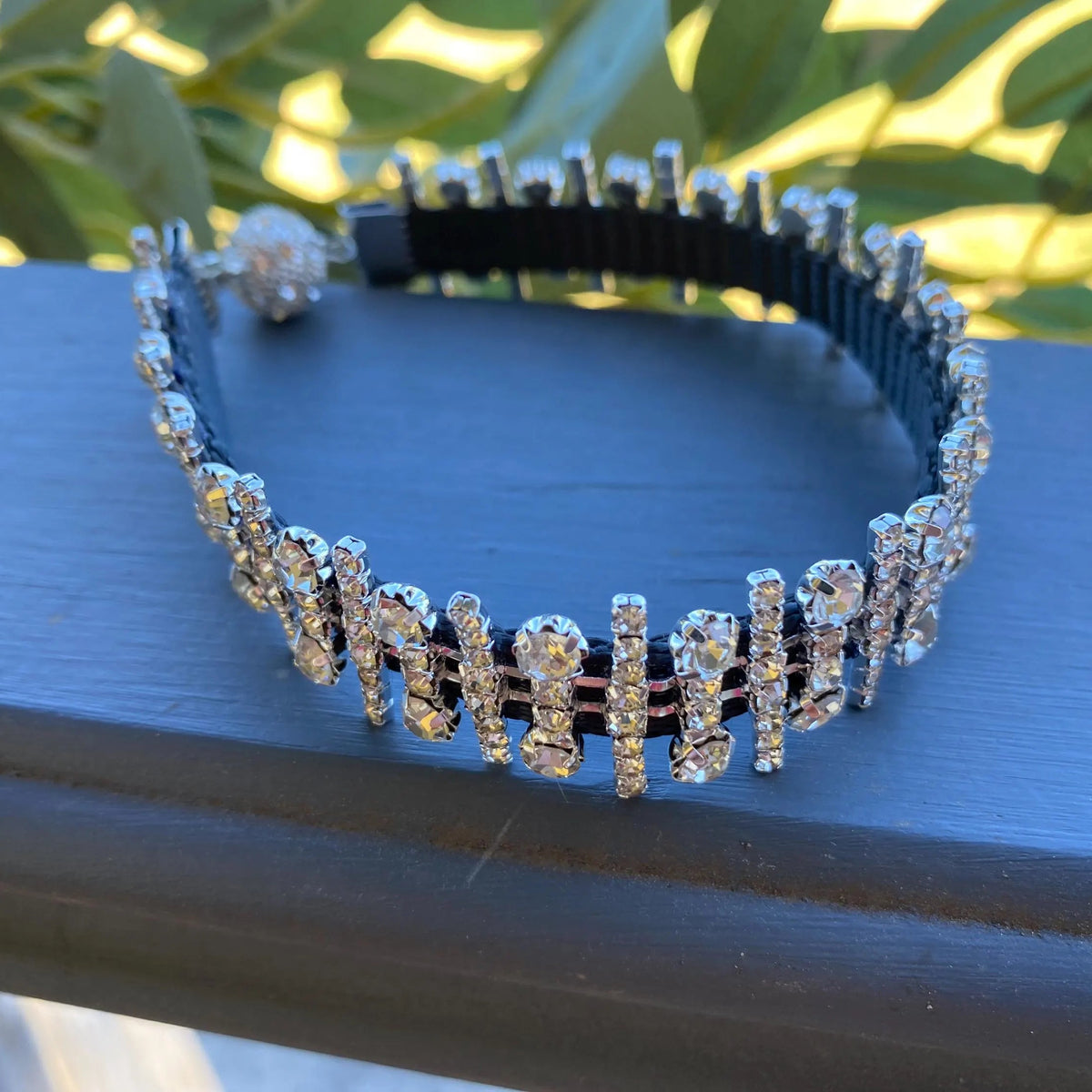 A collar adorned with multiple rows of sparkling clear rhinestones in silver-toned settings, resting on a dark blue wooden surface with green foliage blurred in the background.