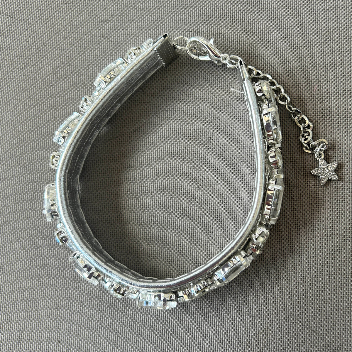 A silver-toned cat collar embellished with clear faceted rhinestones, featuring a lobster clasp, extension chain, and dangling star charm, displayed on a textured gray fabric background.