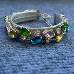 A silver-toned collar adorned with faceted marquise and oval gemstones in green, blue, yellow, and pink, resting on a textured blue fabric background.