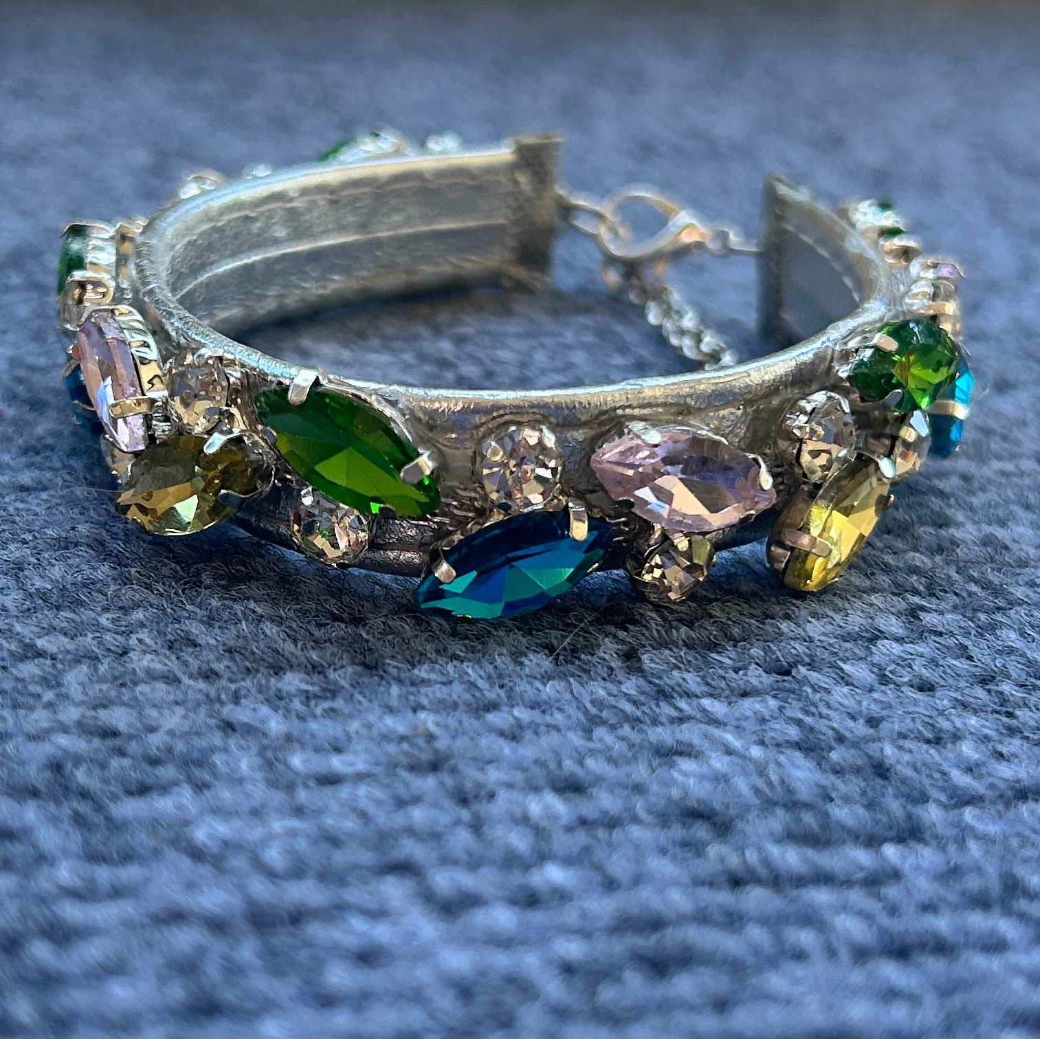 A silver-toned collar adorned with faceted marquise and oval gemstones in green, blue, yellow, and pink, resting on a textured blue fabric background.