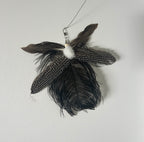 A hanging feather toy with multiple dark brown, black, and white speckled feathers, a fluffy white pom-pom, and a silver metal clasp at the top, photographed against a plain light gray wall.