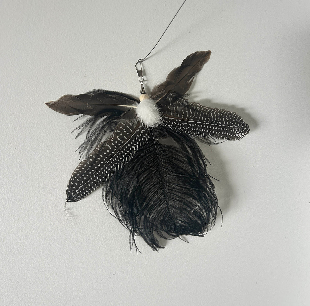 A hanging feather toy with multiple dark brown, black, and white speckled feathers, a fluffy white pom-pom, and a silver metal clasp at the top, photographed against a plain light gray wall.