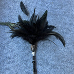 A black feather duster with a metallic silver handle and decorative bead accents, lying on a textured blue-gray fabric background.