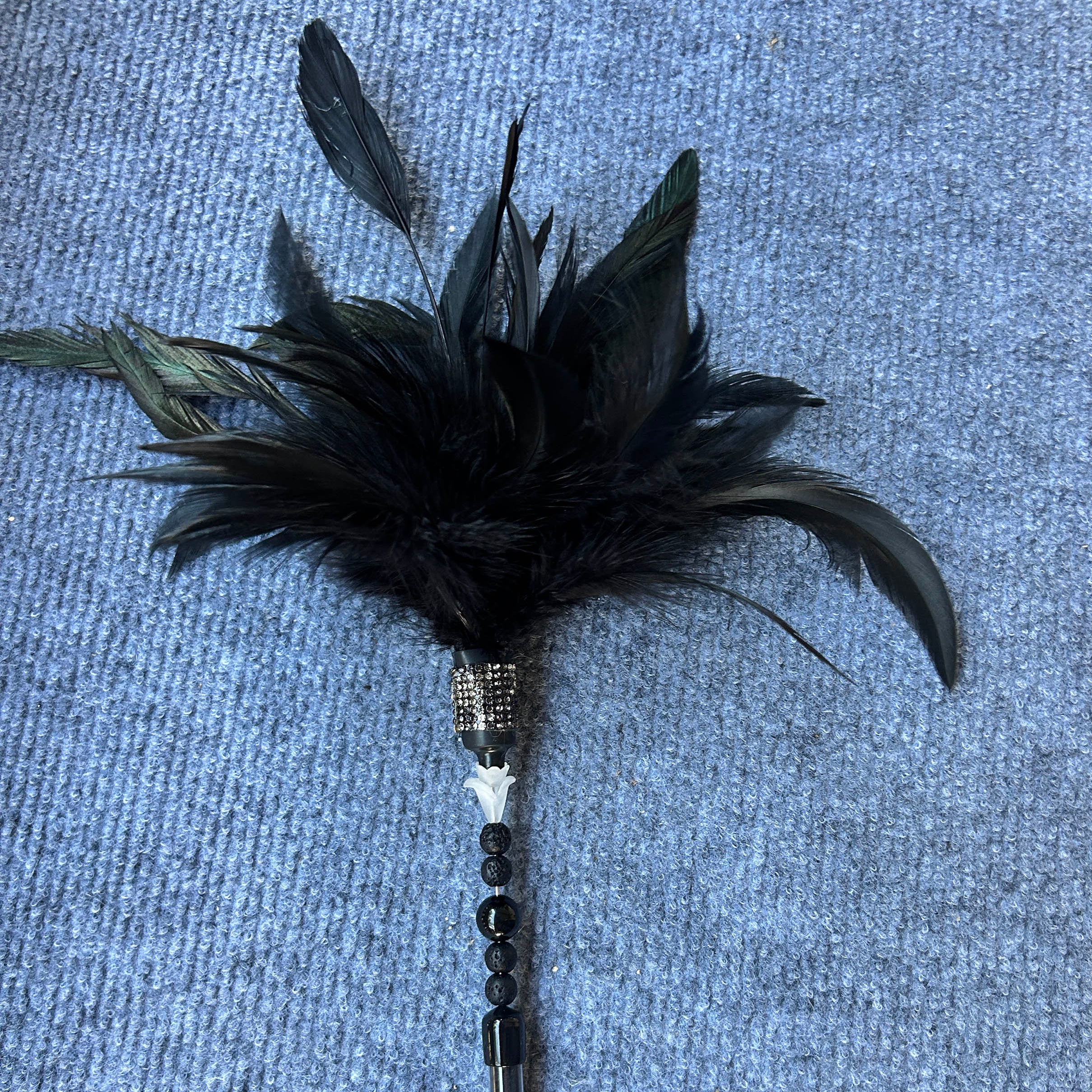 A black feather duster with a metallic silver handle and decorative bead accents, lying on a textured blue-gray fabric background.