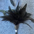 A black feather wand with a rhinestone-embellished silver cap, a white bone-shaped bead, and a black beaded handle, resting on a blue textured carpet.