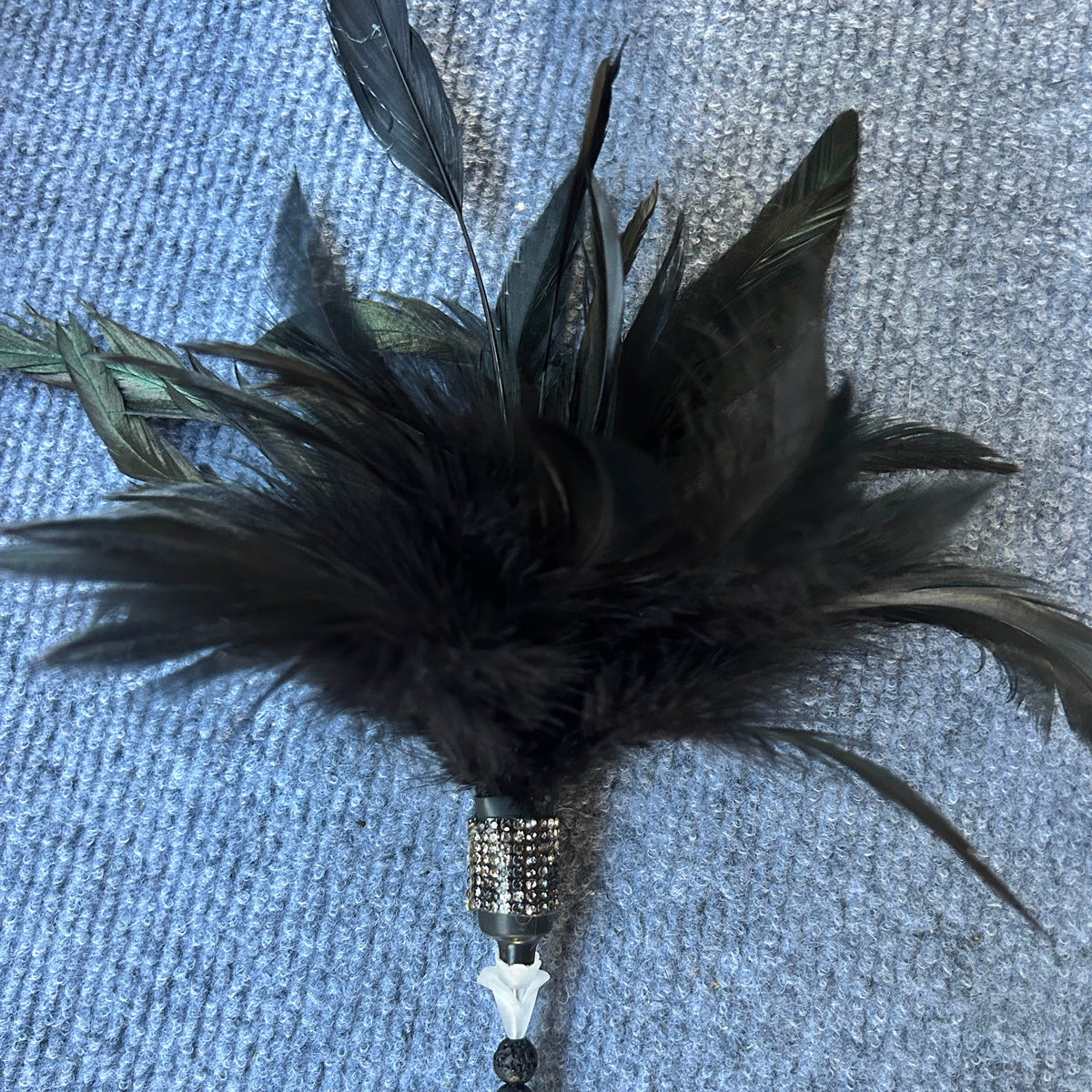 A black feather wand with a rhinestone-embellished silver cap, a white bone-shaped bead, and a black beaded handle, resting on a blue textured carpet.