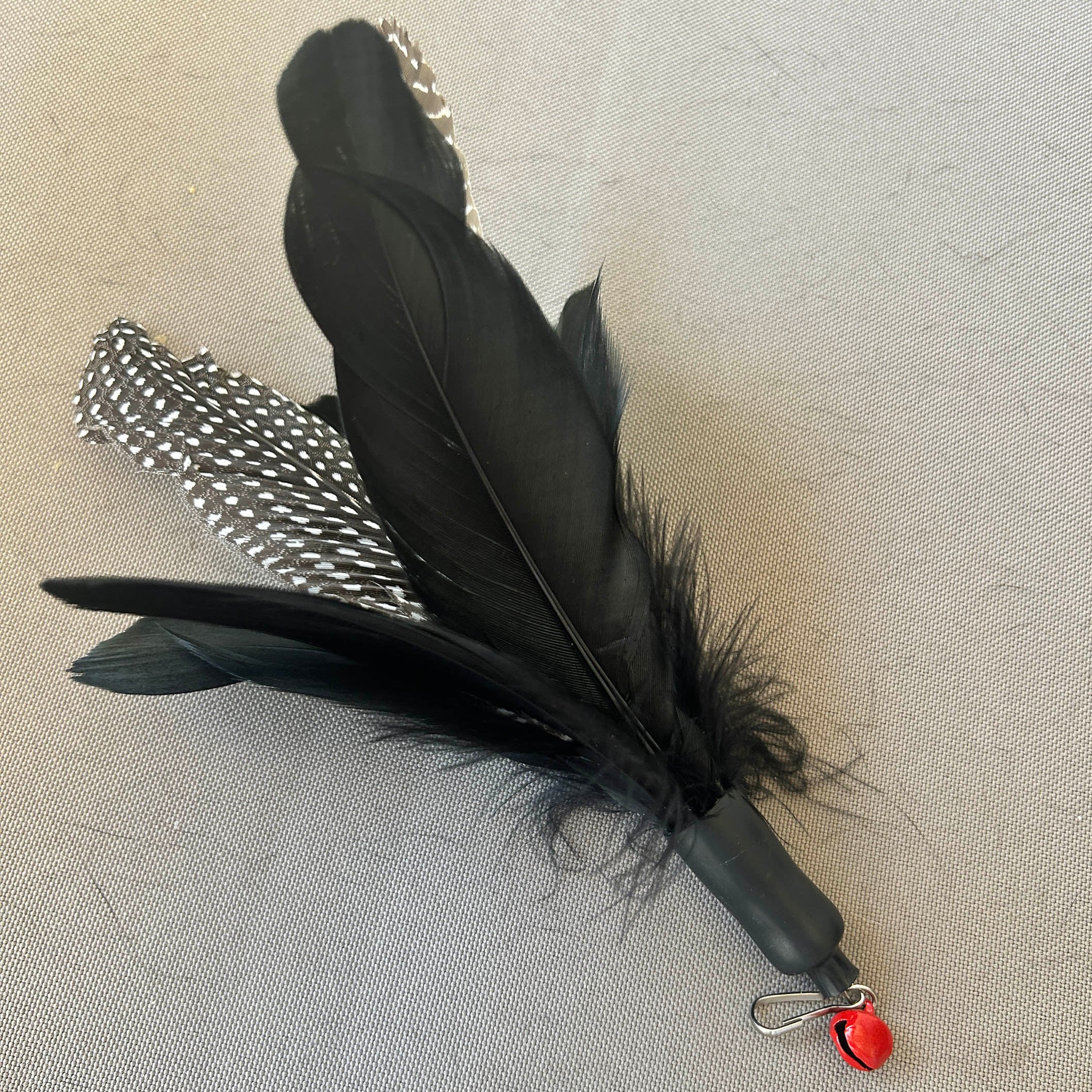 A black feather wand toy with two black and white speckled feathers, a black handle, and a small red bell at the end, resting on a light beige textured fabric background.