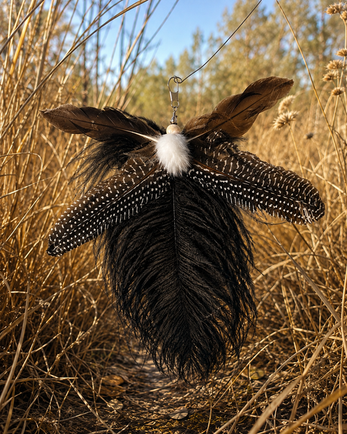 A feather ornament cat toy attachment featuring a large black ostrich feather, a black-and-white barred feather, and a brown feather, set against a background of dry grass and trees.