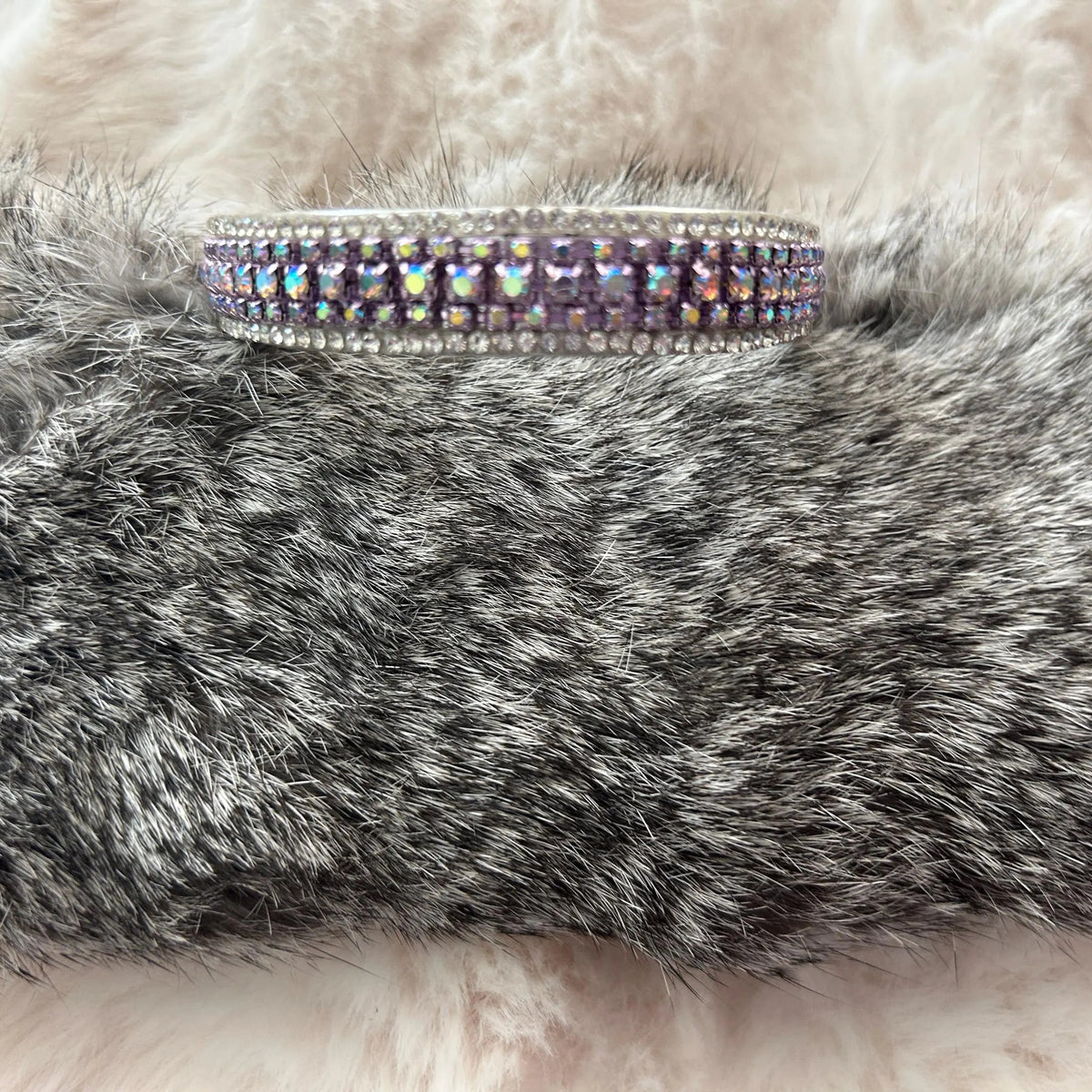 A silver-toned double-row cuff bracelet with iridescent purple and blue rhinestones, displayed on a gray faux fur surface against a white faux fur background.
