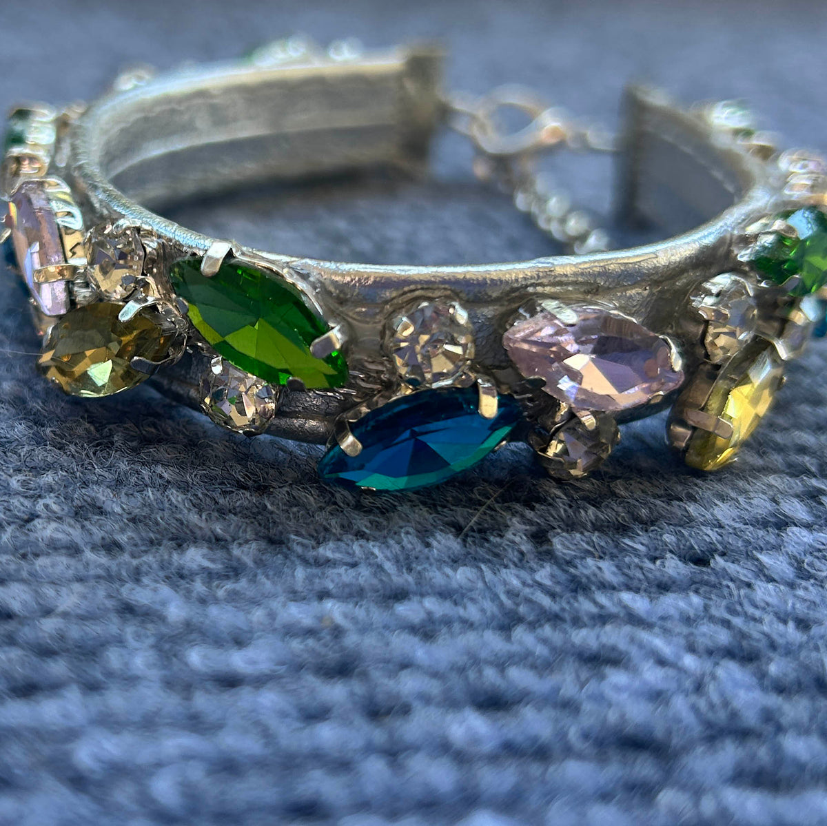 A vintage-style silver collar adorned with multiple colorful faceted rhinestones in green, blue, yellow, and pink, resting on a textured gray fabric background.