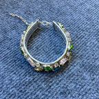 A silver-toned collar adorned with baguette and round crystals in green, pink, and blue, displayed on a textured blue fabric background.