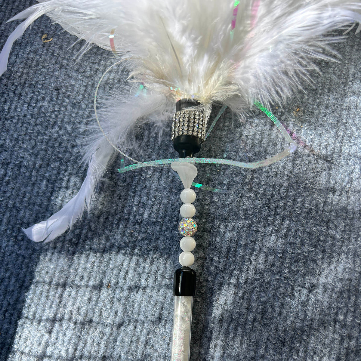 A white feather wand with white beads, a rhinestone-embellished black cap, and a white handle, lying on a gray textured blanket.