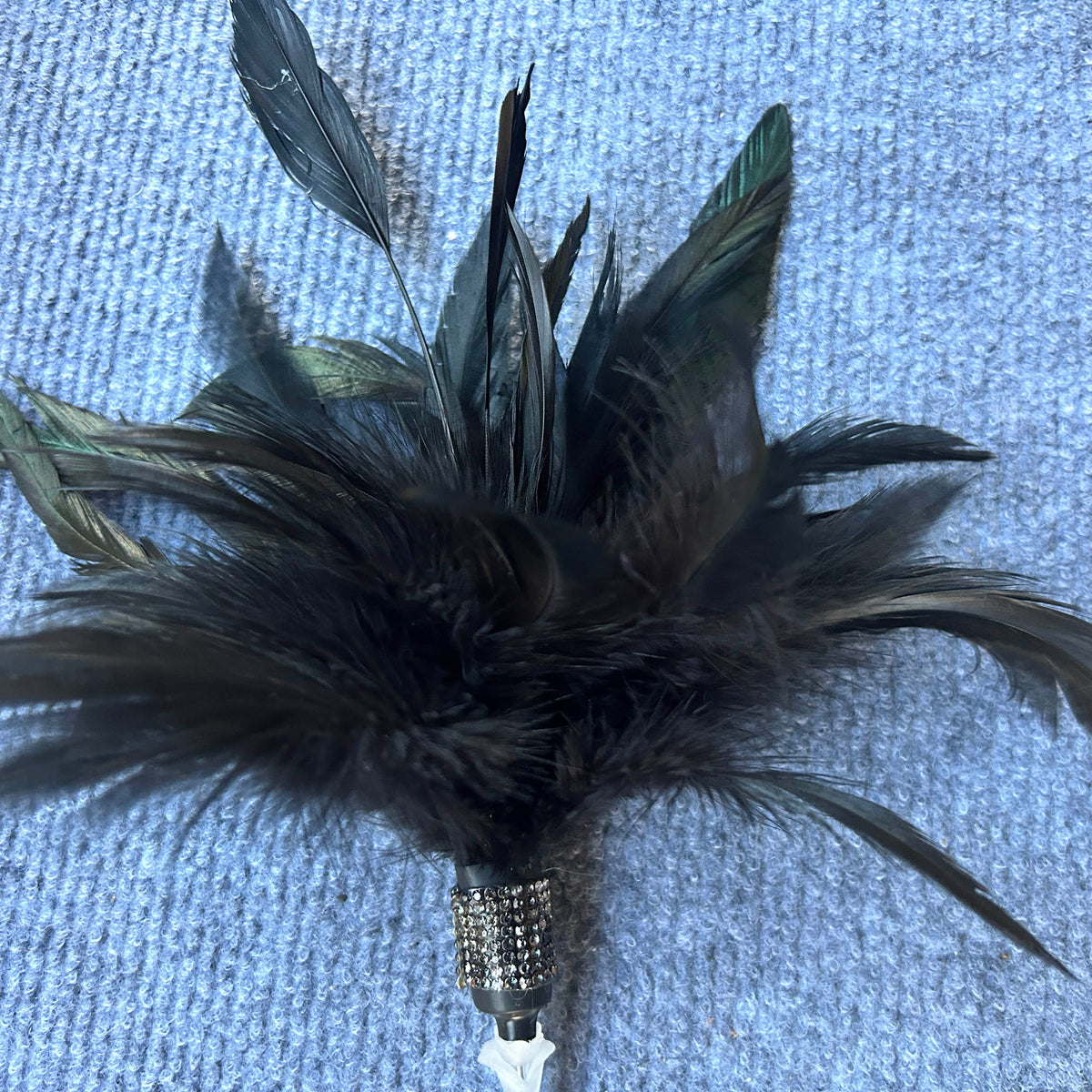 A decorative feather ornament with multiple black and dark gray feathers attached to a rhinestone-embellished silver base, photographed on a textured light blue fabric background.