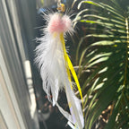 A hanging feather toy with a fluffy pink top, long white feathers, and a bright yellow ribbon tail, photographed outdoors against a background of green leaves and sunlight.
