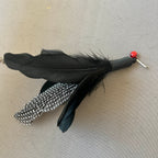 A black feather cat lure with a black and white speckled feather, a black body, and a small red bead accent, shown on a neutral beige textured background.
