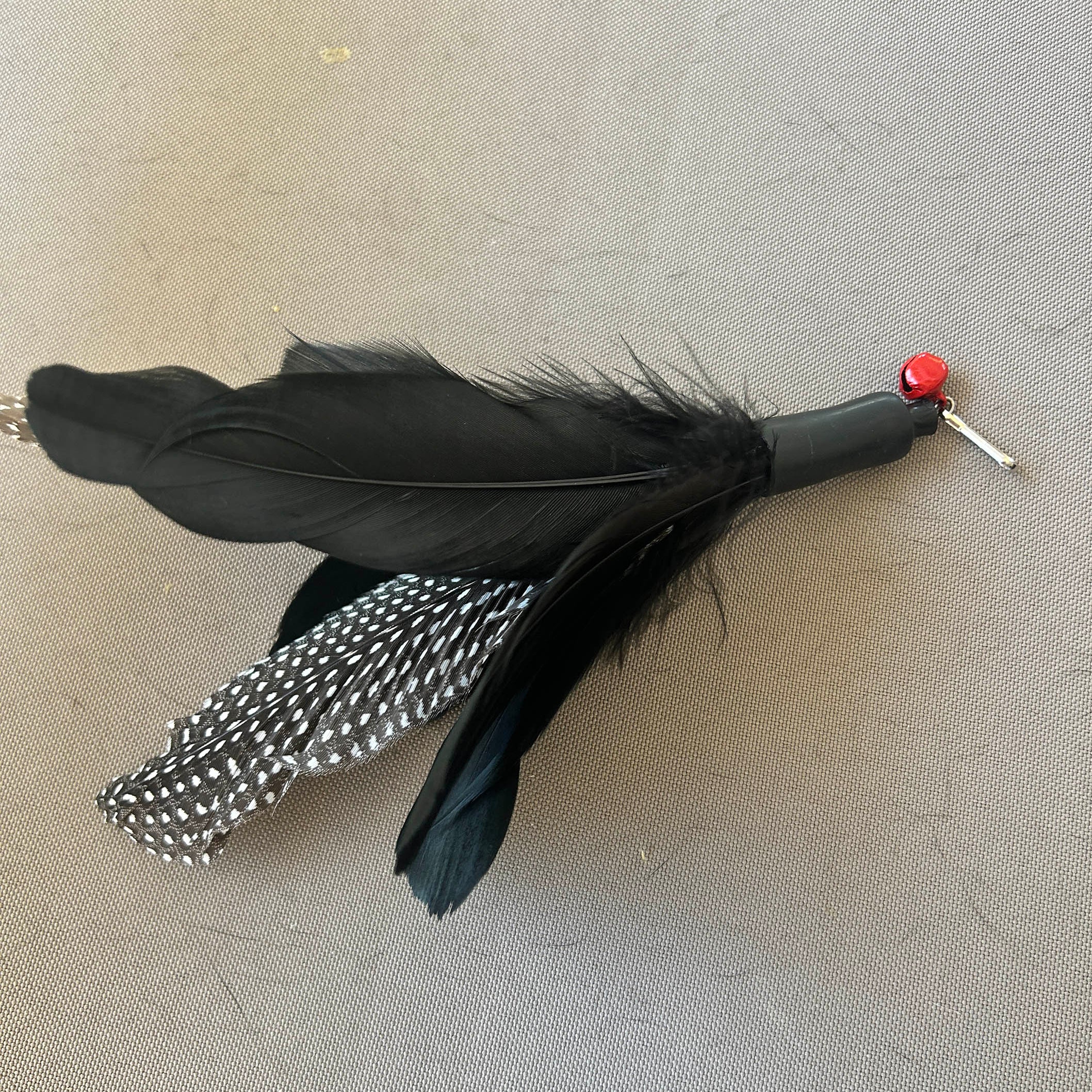 A black feather cat lure with a black and white speckled feather, a black body, and a small red bead accent, shown on a neutral beige textured background.