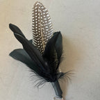 A black feather cat  lure with a black-and-white speckled patterned feather, black feathers, and a small red bead at the tip, shown on a neutral gray textured background.