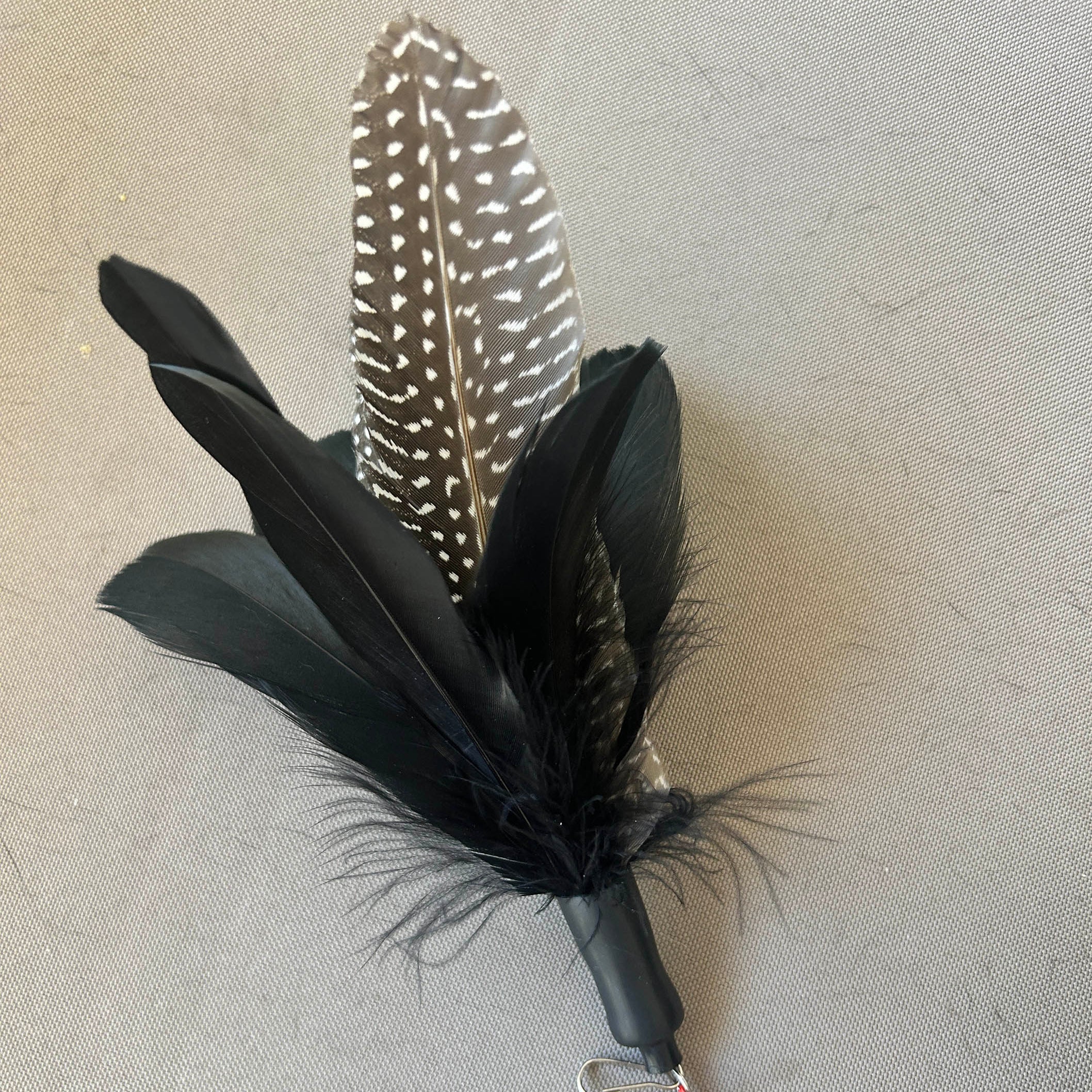 A black feather cat  lure with a black-and-white speckled patterned feather, black feathers, and a small red bead at the tip, shown on a neutral gray textured background.