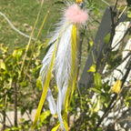 A hanging feather toy with white feathers, yellow ribbons, and a pink fluffy pom-pom, photographed outdoors among green plants and branches.