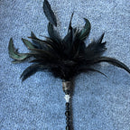 A black feather duster wand with iridescent green feathers, a beaded handle, and a rhinestone-embellished ferrule, lying on a textured blue-gray fabric background.
