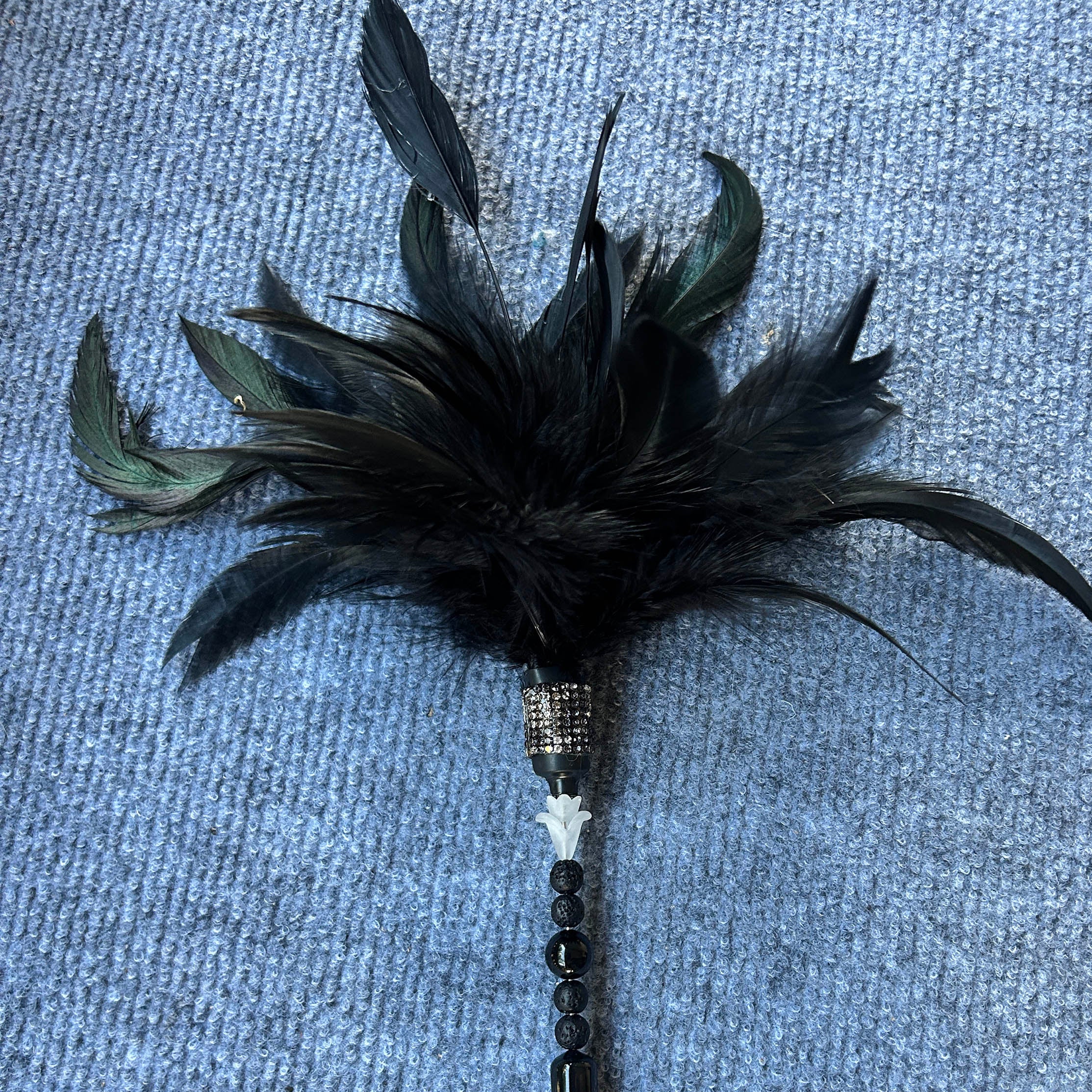 A black feather duster wand with iridescent green feathers, a beaded handle, and a rhinestone-embellished ferrule, lying on a textured blue-gray fabric background.