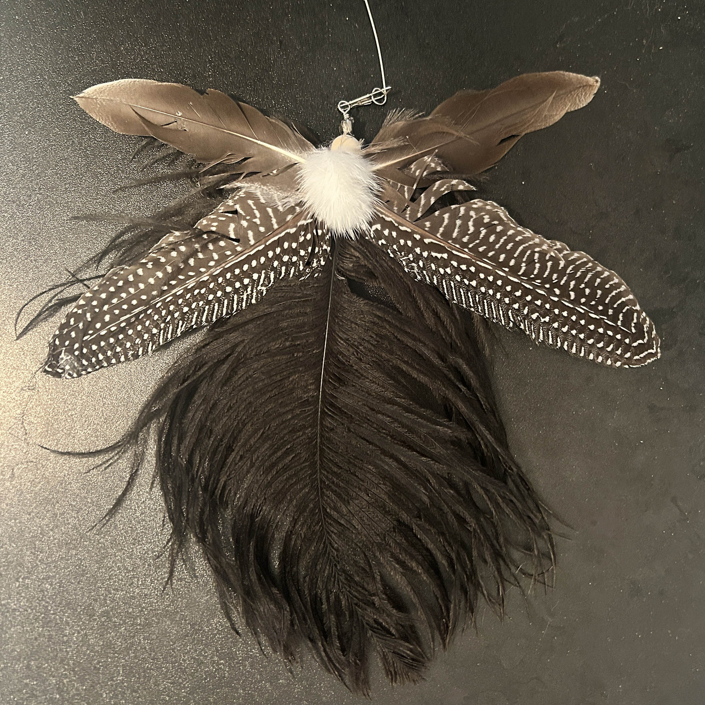 A cat toy made from multiple natural feathers, including a large dark brown feather, lighter brown feathers, white speckled feathers, and a fluffy white center, photographed against a dark textured background.