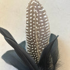 A decorative feather arrangement featuring a tall brown and white spotted central feather with two black feathers layered around it, set against a light beige fabric background.