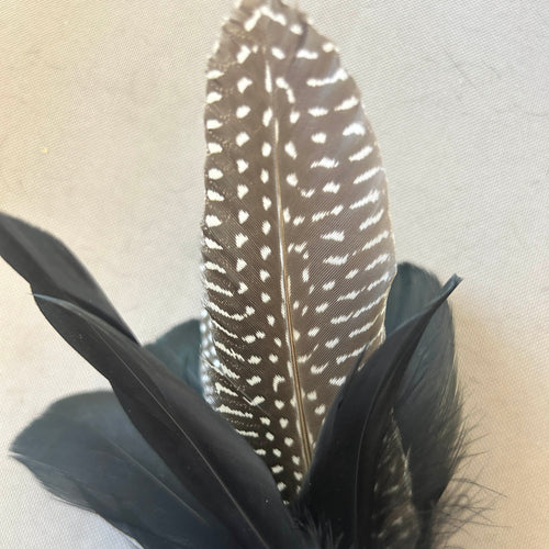 A decorative feather arrangement featuring a tall brown and white spotted central feather with two black feathers layered around it, set against a light beige fabric background.