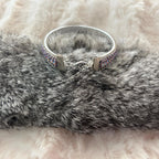 A silver-toned cuff bracelet with iridescent purple and blue rhinestone detailing, displayed on a gray faux fur surface with a soft white faux fur background.