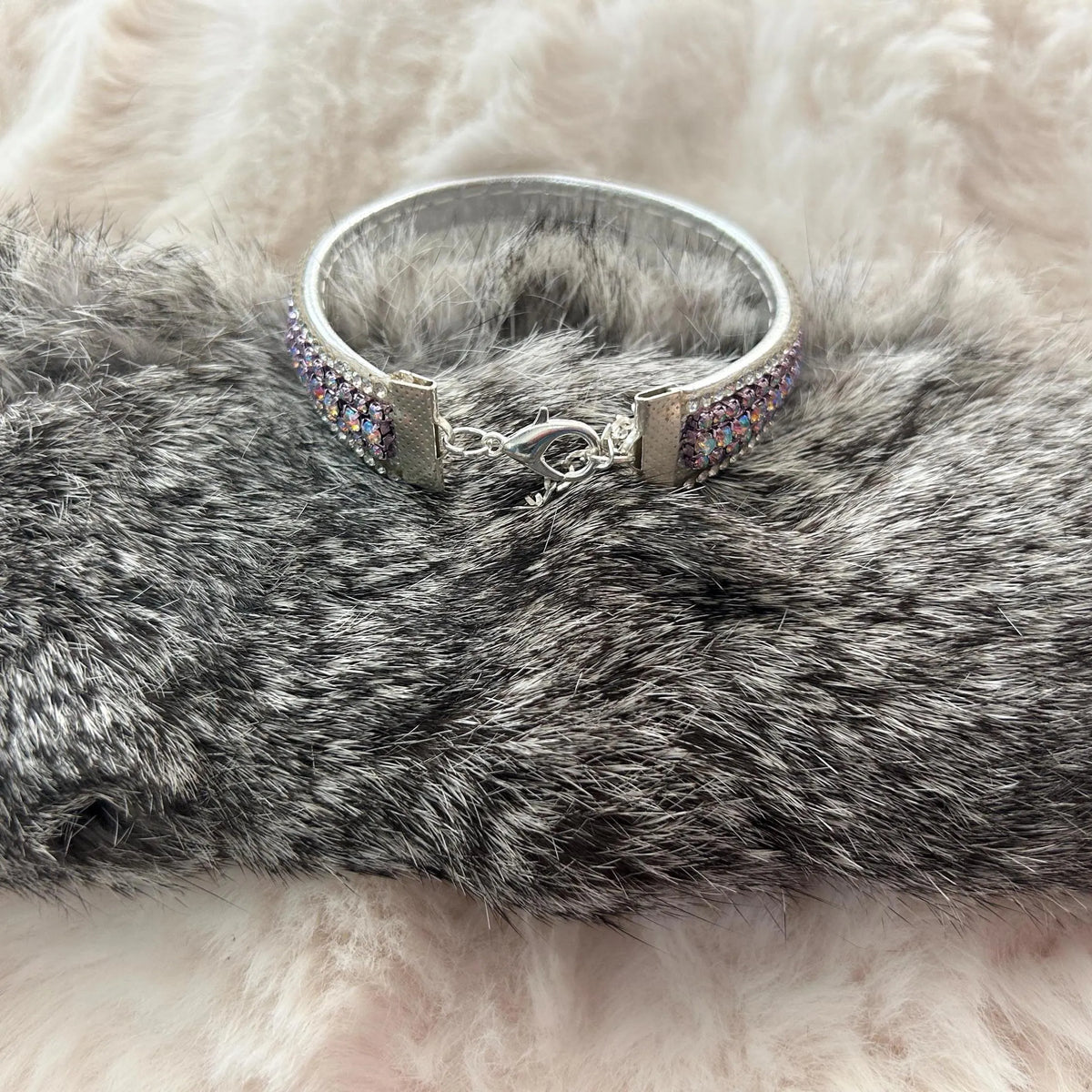 A silver-toned cuff bracelet with iridescent purple and blue rhinestone detailing, displayed on a gray faux fur surface with a soft white faux fur background.
