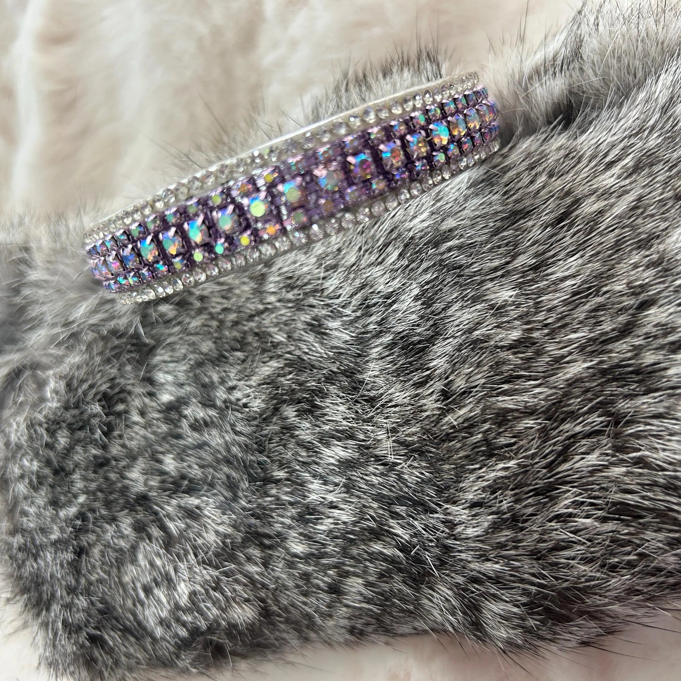 A close-up of a gray faux fur sleeve or cuff with a wide silver-toned band embellished with iridescent rhinestones, resting on a soft cream-colored faux fur surface.