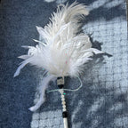 A white ostrich feather duster with a beaded handle, laid on a blue-gray textured blanket with dappled sunlight and shadows.