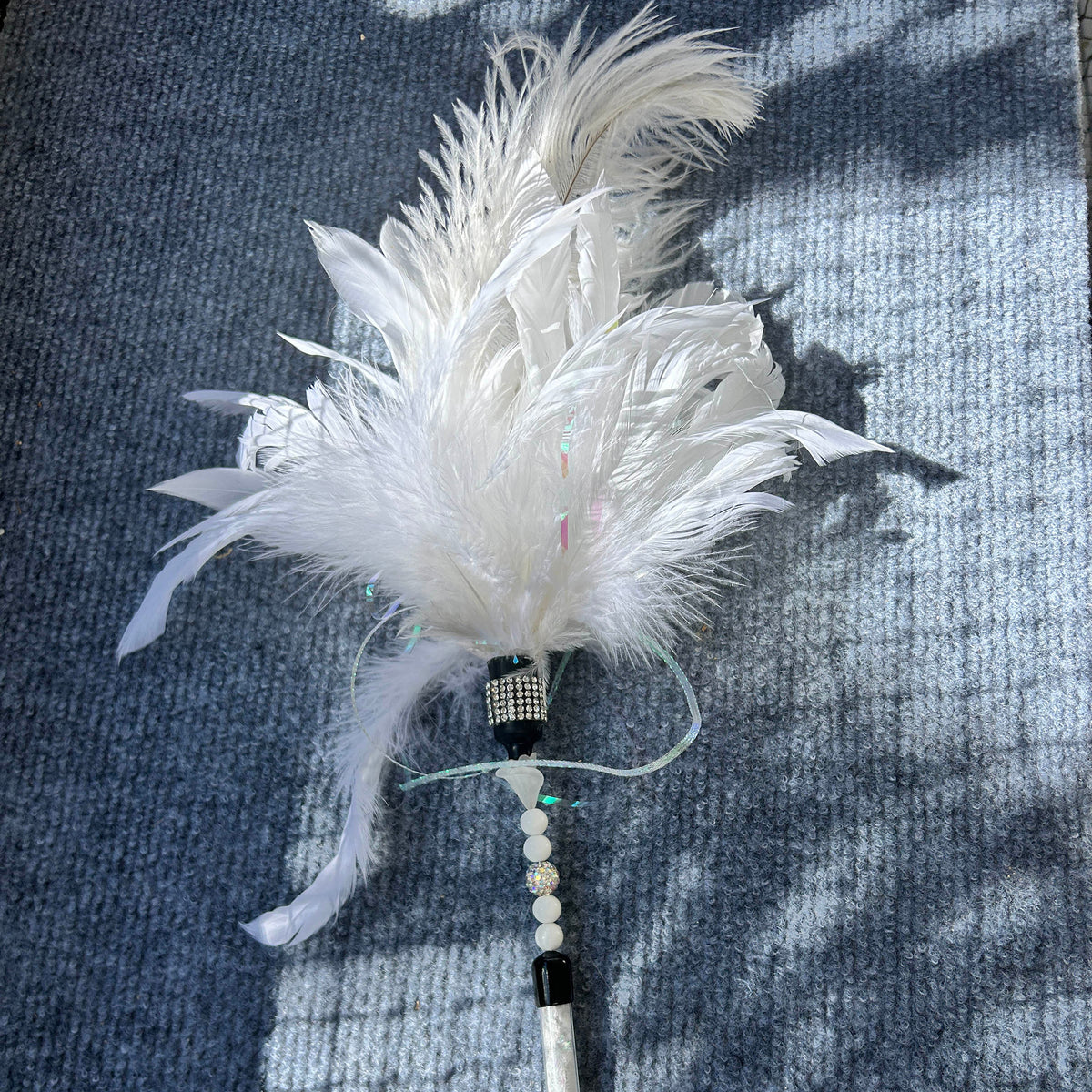 A white ostrich feather duster with a beaded handle, laid on a blue-gray textured blanket with dappled sunlight and shadows.