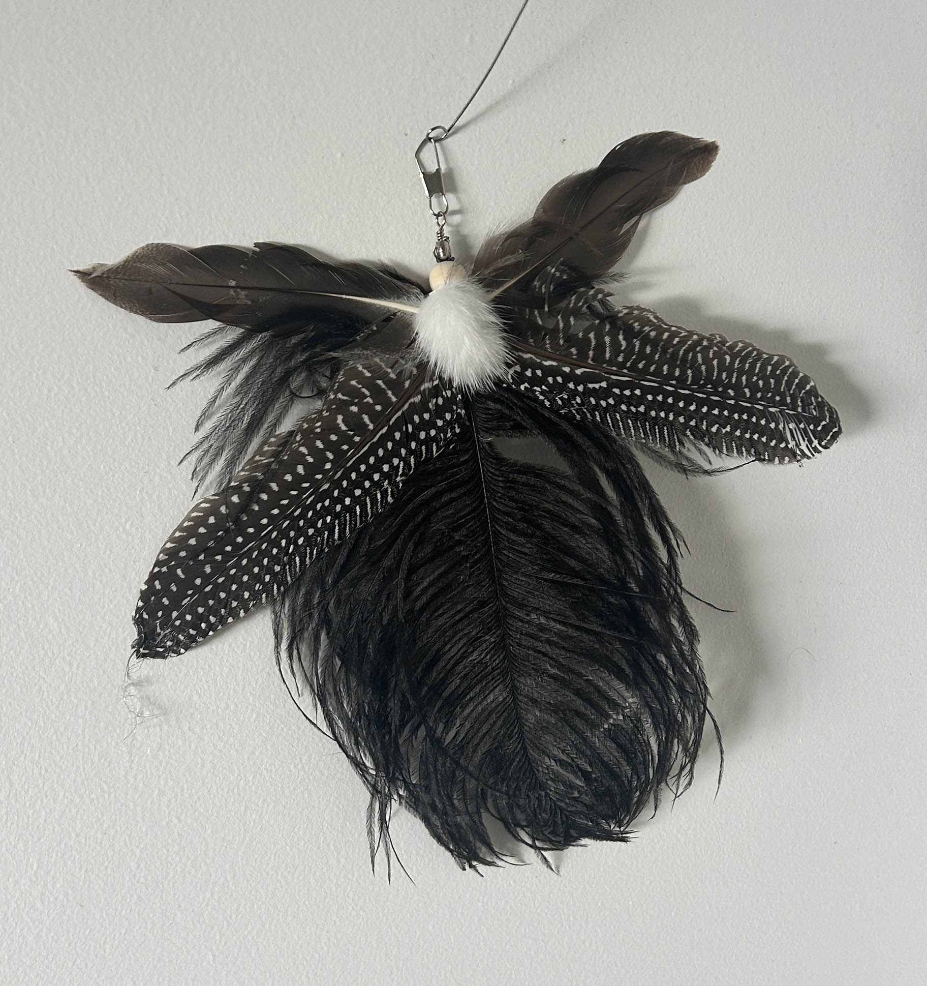 A  feather cat toy made from several dark feathers, including black and brown feathers with white speckles, a white fluffy centre, and a metal hanging loop.