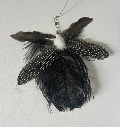 A  feather cat toy made from several dark feathers, including black and brown feathers with white speckles, a white fluffy centre, and a metal hanging loop.