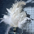 A white decorative feather ornament with layered soft feathers, a rhinestone-embellished black bead, iridescent ribbon, and white beads, photographed on a dark textured fabric background.