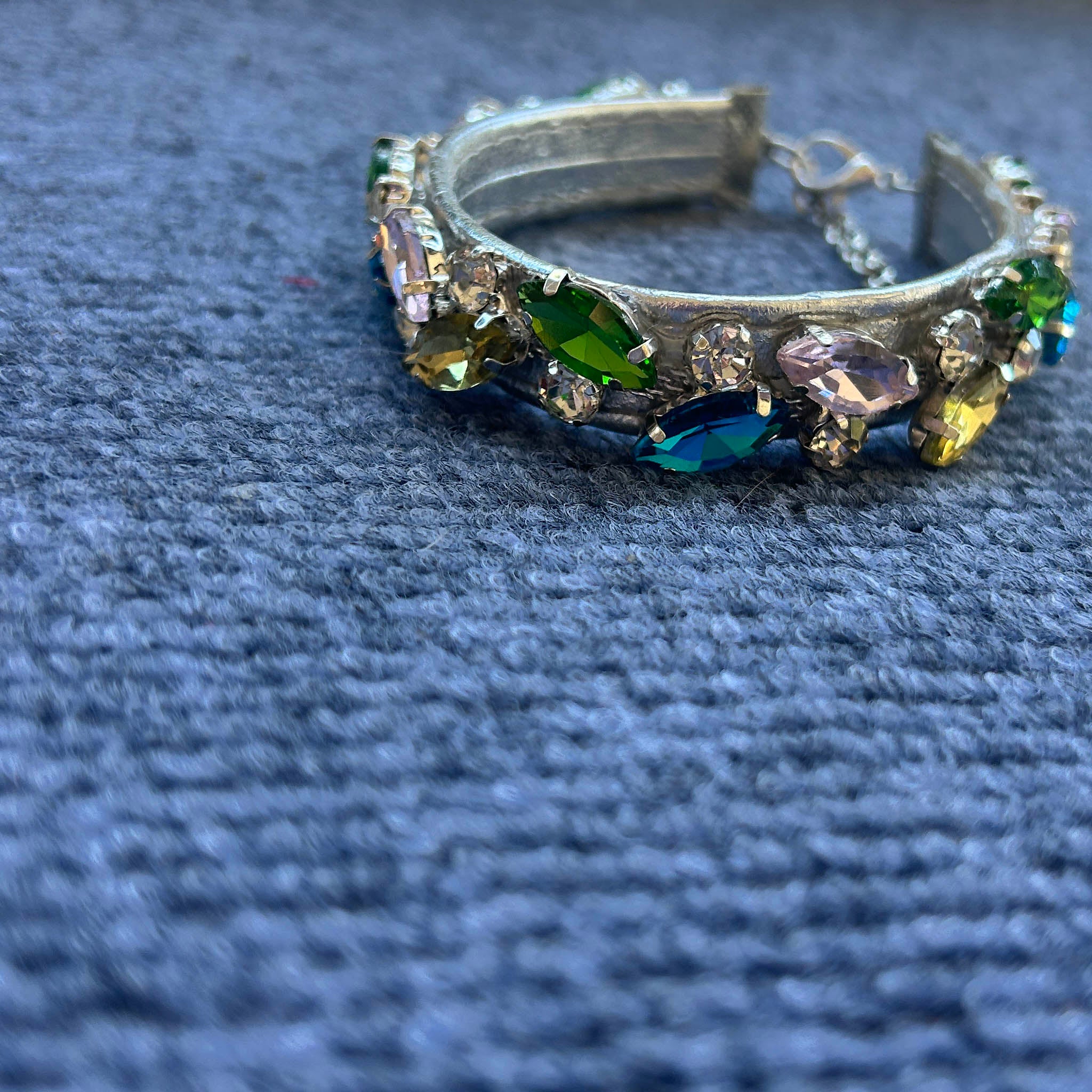 A silver collar adorned with colorful faceted rhinestones in green, blue, yellow, and pink, resting on a textured blue fabric background.