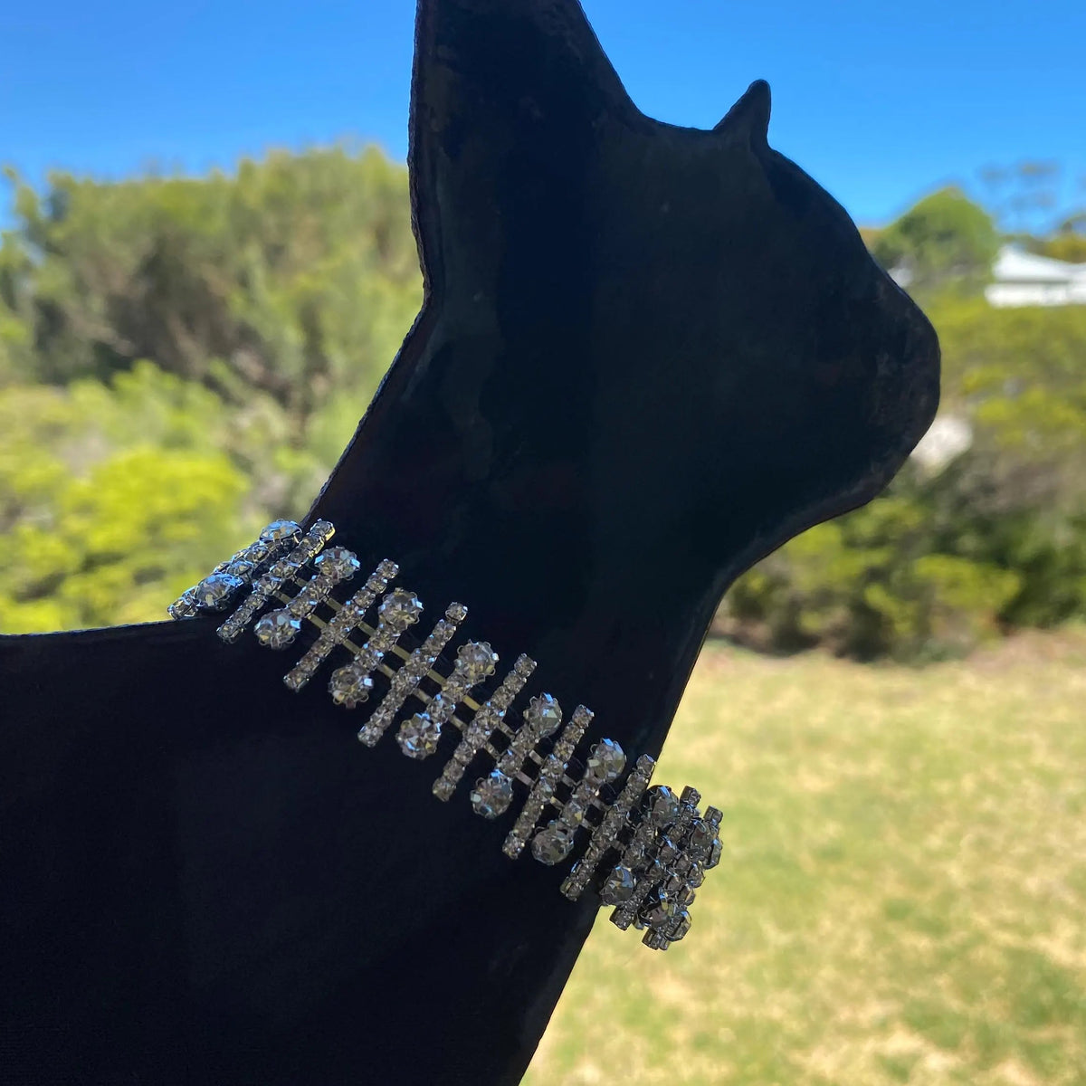 A close-up of a black cat statue wearing a sparkling silver rhinestone collar, set against a blurred outdoor background of green trees and blue sky.