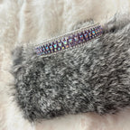 A silver-toned cuff bracelet embellished with rows of iridescent purple and blue rhinestones, resting on a textured gray faux fur surface with a soft white background.