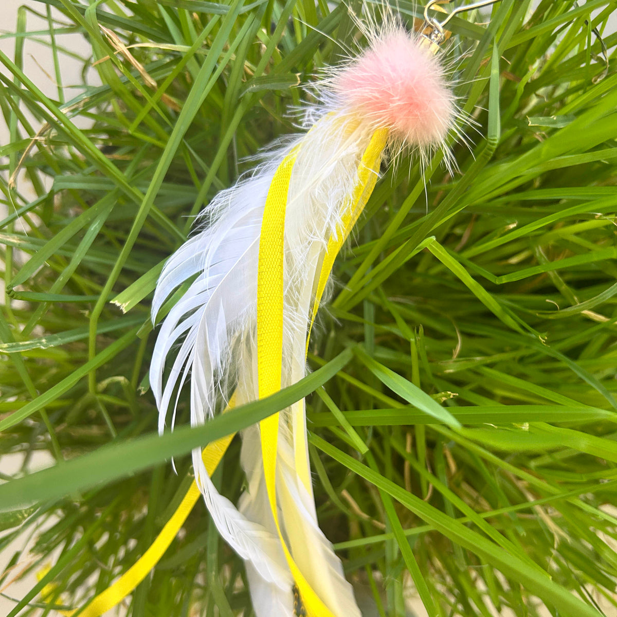 A feather toy with a white feather, a yellow ribbon, and a pink fuzzy pom-pom, shown hanging in green grass.