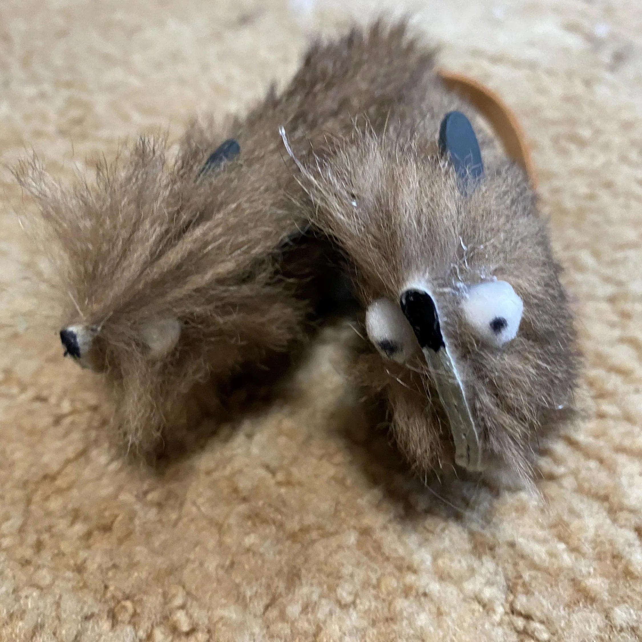 Two small brown furry toy mice with white googly eyes and black noses, lying on a beige carpeted floor.