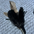 A cat lure with black feathers, a speckled brown and white feather, a black body, and a small red bead on a textured gray background.