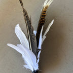 Three decorative feathers attached to a black stem, featuring white feathers at the base and two long brown feathers with white tips, displayed on a neutral beige background.