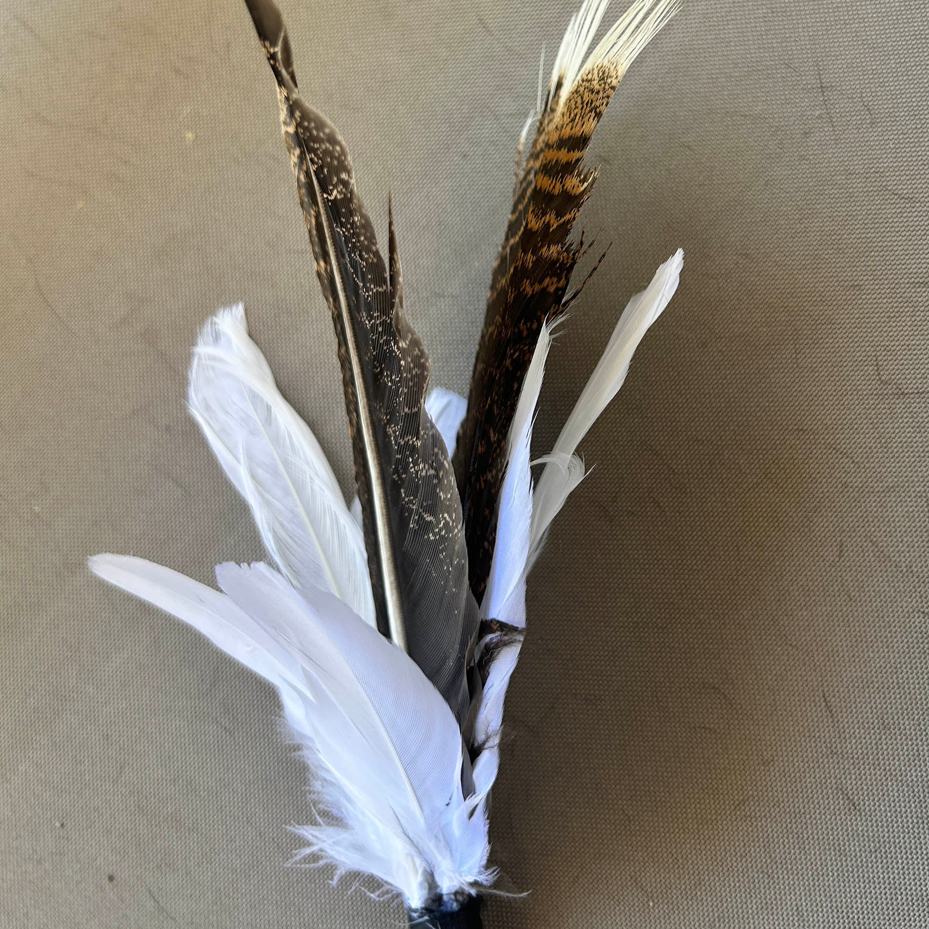 Three decorative feathers attached to a black stem, featuring white feathers at the base and two long brown feathers with white tips, displayed on a neutral beige background.