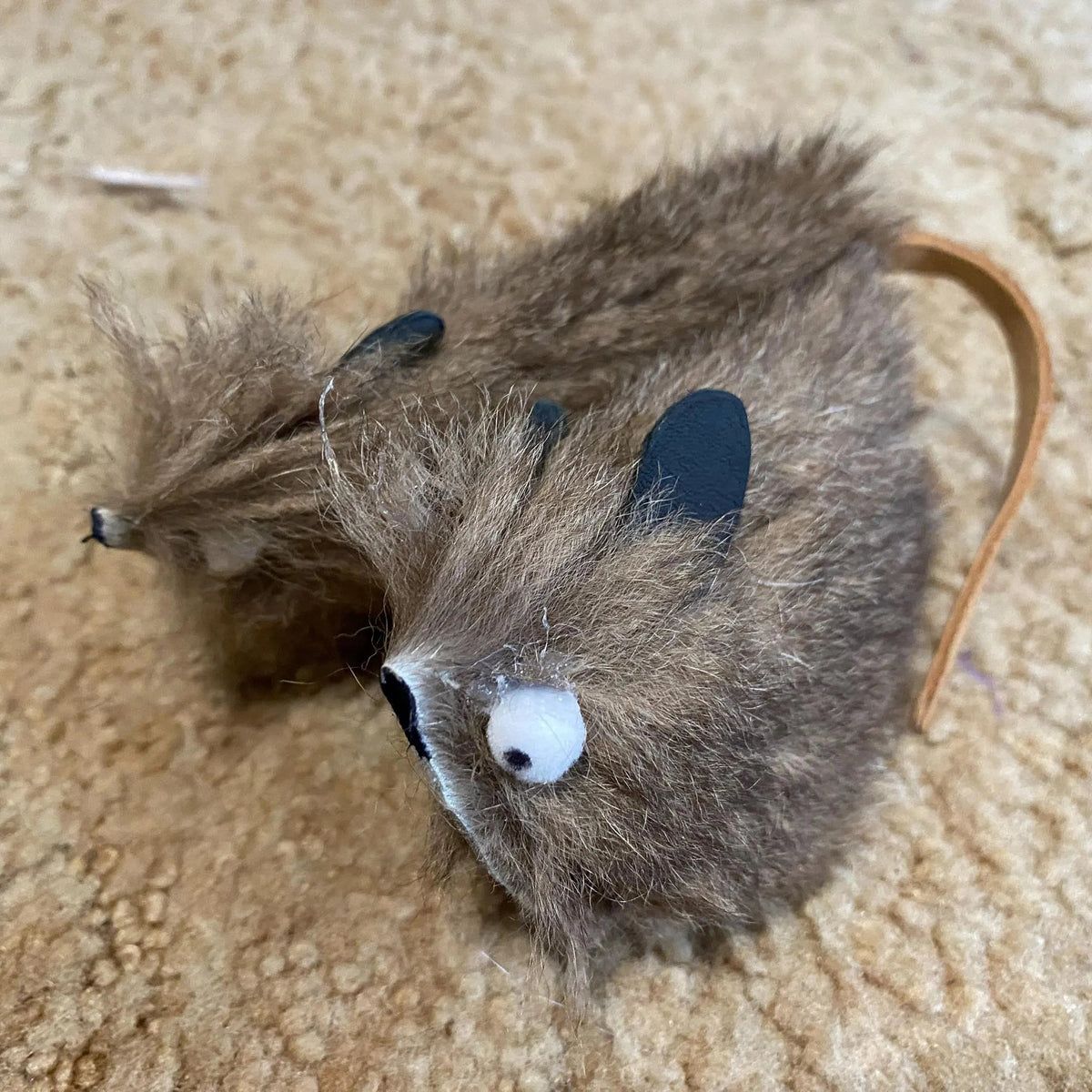 A brown faux-fur toy mouse with a long tan tail, black ears, and a white eye, lying on a beige carpeted floor.